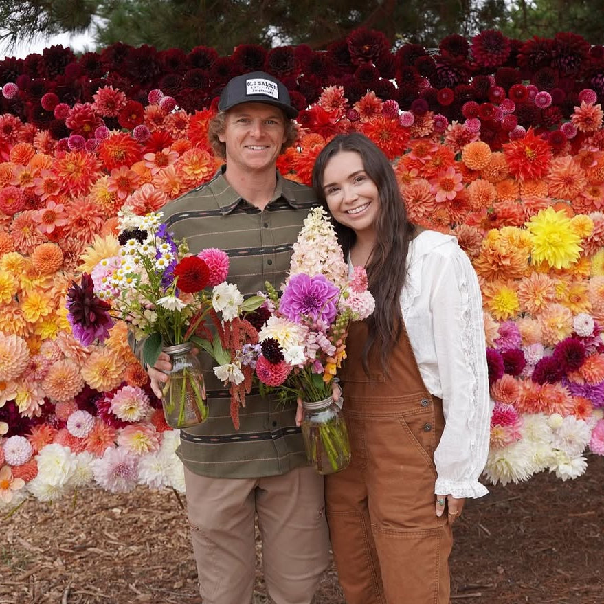 Two people standing in front of a vibrant flower display with trees in the background.