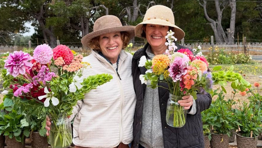 Two women holding flower bouquets in a garden setting with trees and plants around.