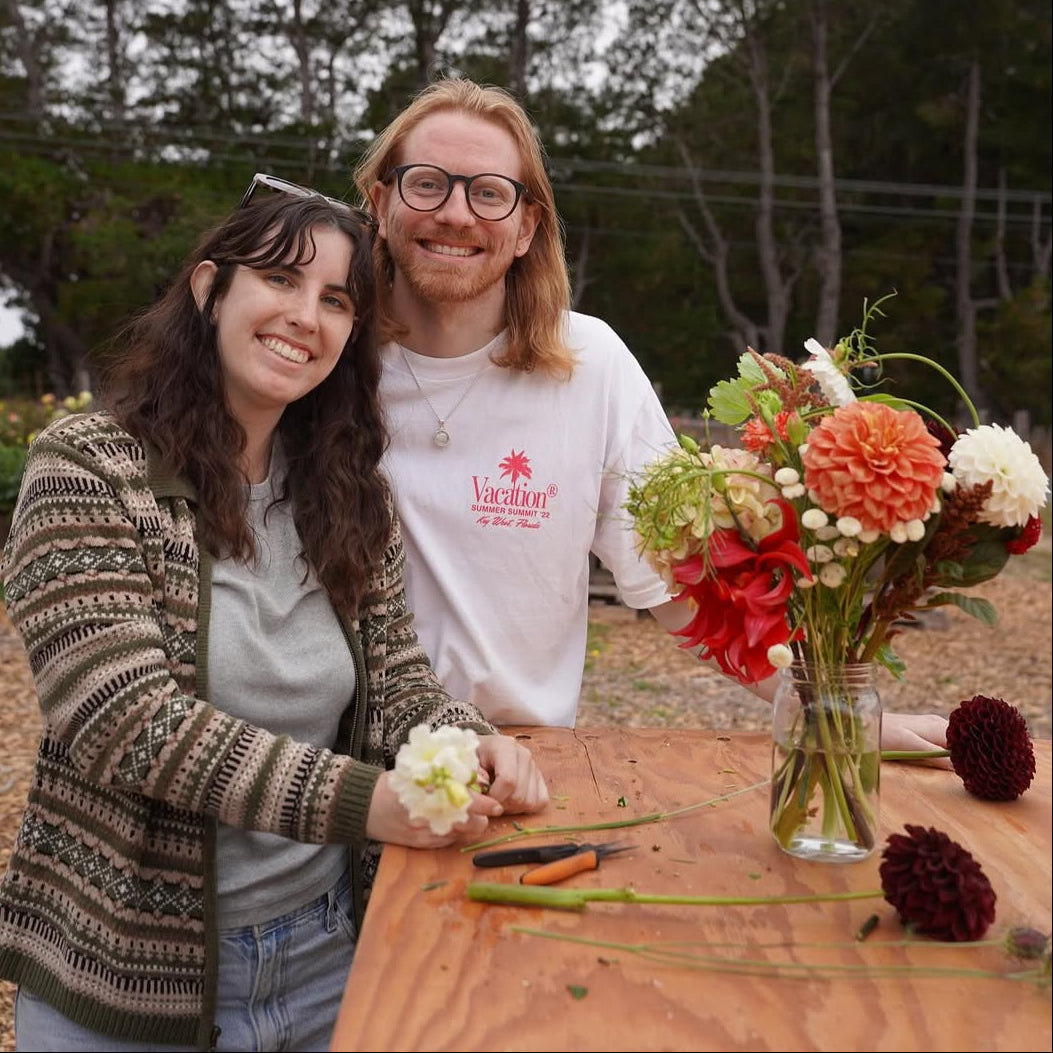 Two people standing behind a table with flowers and tools outdoors.
